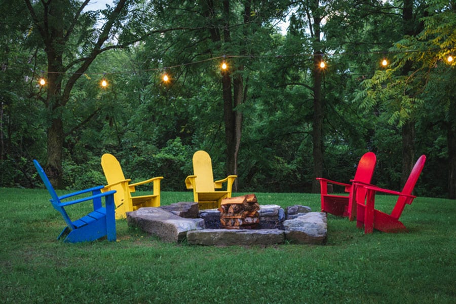 Five colorful Adirondack chairs surround a stone fire pit on a grassy lawn, with string lights overhead and healthy trees in the background, their lush foliage thriving thanks to regular tree trimming. Five colorful Adirondack chairs surround a stone fire pit on a grassy lawn, with string lights overhead and healthy trees in the background, their lush foliage thriving thanks to regular tree trimming.