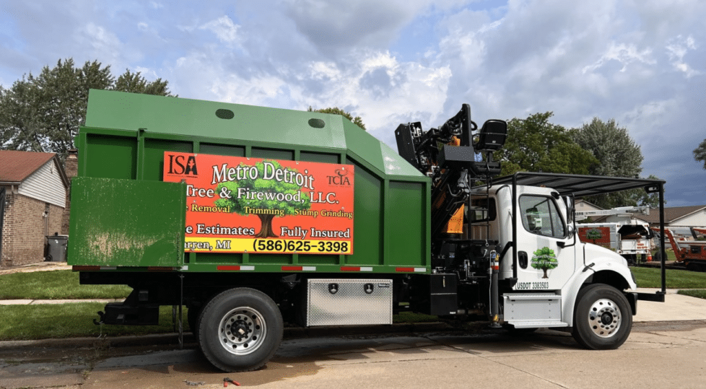 A green and white Metro Detroit Tree & Firewood truck is parked on a residential street in New Haven MI. The truck displays Tree Service details and carries equipment used for tree care. Houses and trees can be seen in the background.