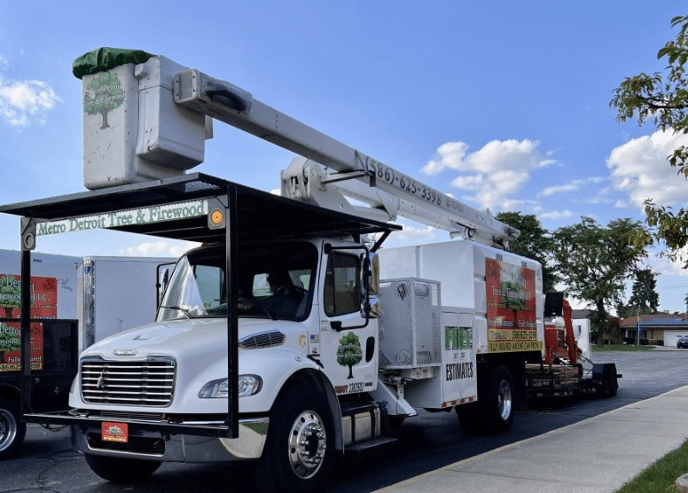 A white Tree Service truck with a bucket lift is parked on a street in New Baltimore, MI. The truck displays Metro Detroit Tree & Firewood and a green tree logo. The sky is clear with just a few clouds.