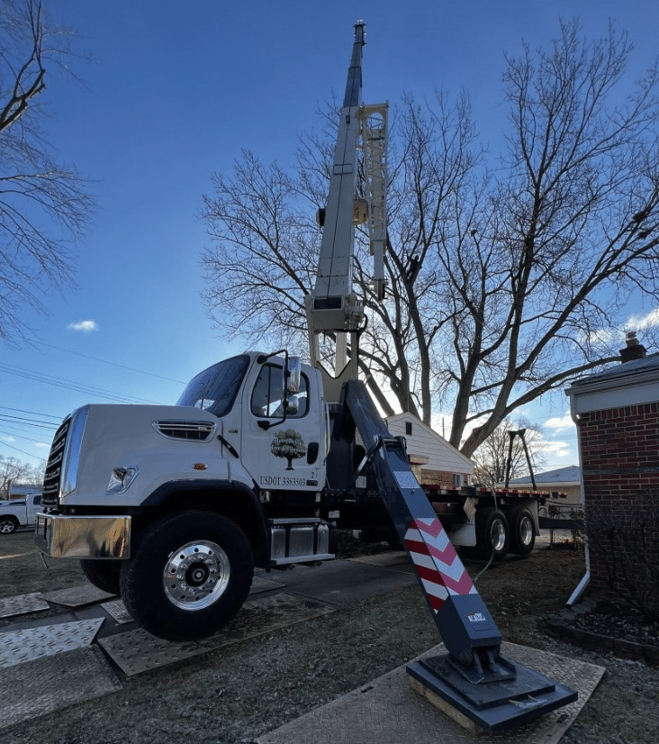 A large white truck-mounted crane from a Tree Service in Harrison Township, MI is parked on a lawn, its extendable boom raised and stabilizing outriggers deployed. Leafless trees and a brick building are visible under a partly cloudy sky.