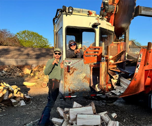 Two men posing next to a tractor equipped with a large circular saw blade, surrounded by piles of split firewood.
